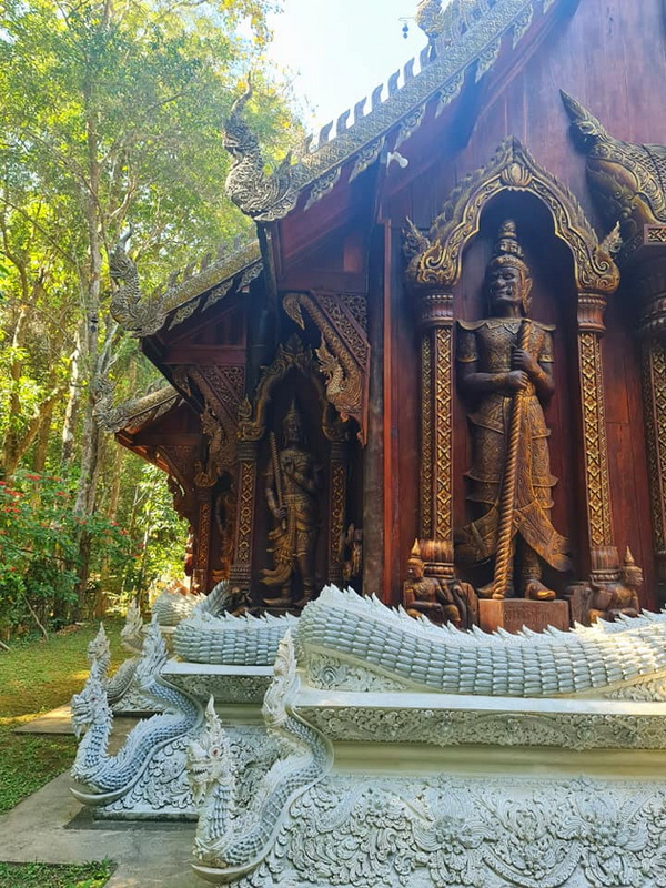 Intricately carved wooden temple with mythical creature sculptures and serpent base, surrounded by greenery at Wat Luang Khun Win.