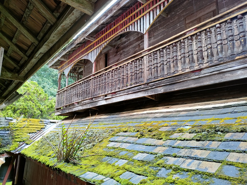 Wooden Building with Mossy Roof at Wat Luang Khun Win Close-up of a wooden building's balcony and mossy roof with lush greenery nearby at Wat Luang Khun Win.
