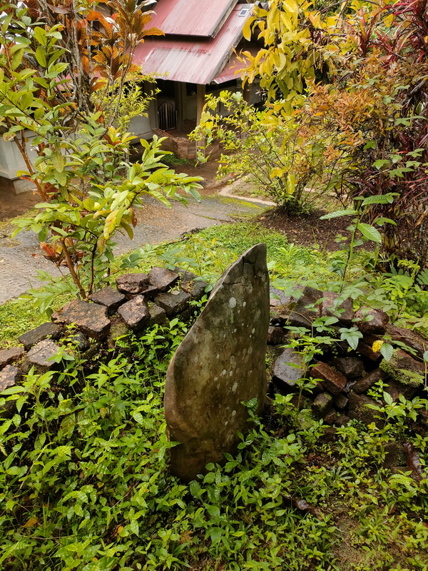 A stone slab surrounded by plants and a brick wall near a house with red roof at Wat Luang Khun Win.