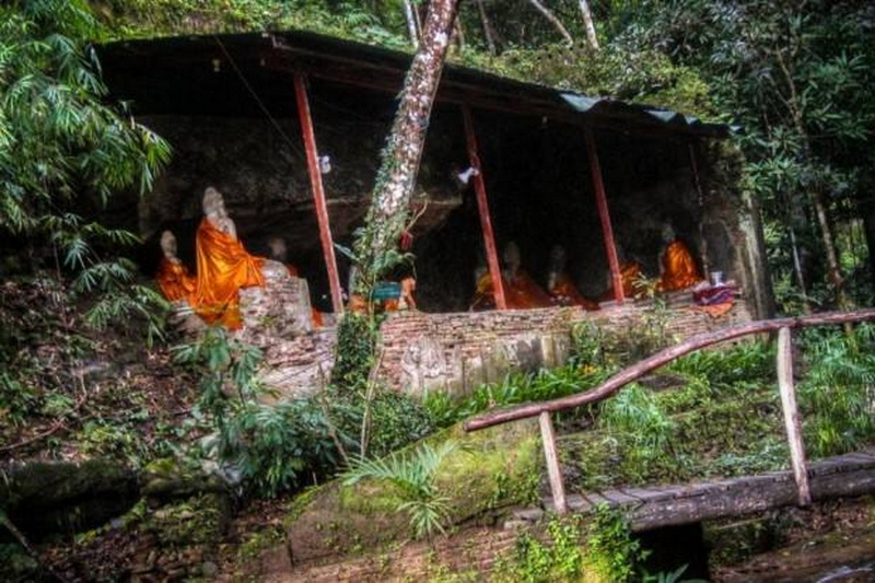 Buddha statues covered with orange cloth under a shaded shelter in a forested area at Wat Pha Lad.