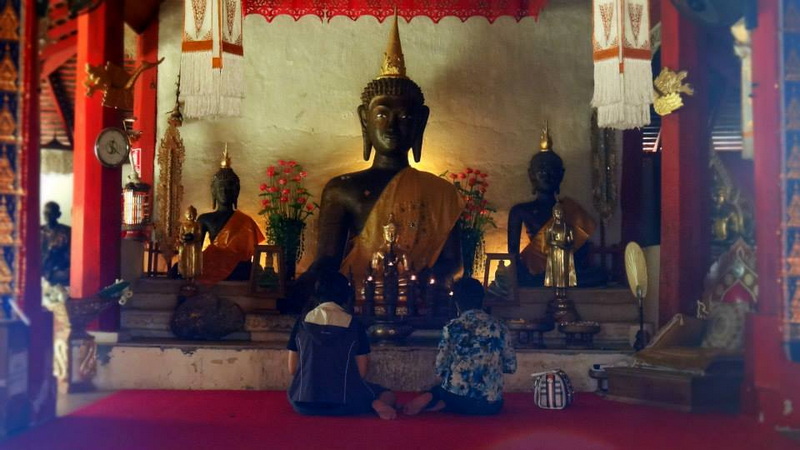 Children sitting in front of Buddha statue inside a richly decorated temple at Wat Pha Lad.