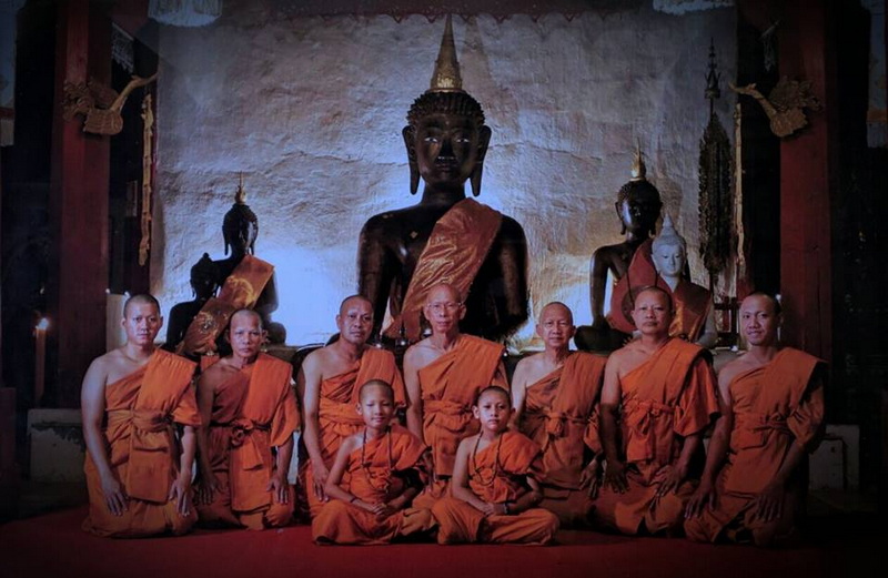 Monks in orange robes sitting in front of large Buddha statues inside a temple at Wat Pha Lad.