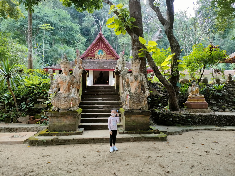 Person standing in front of carved statues and stairs leading to a temple in a lush forest at Wat Pha Lad.