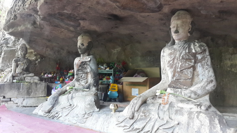 Stone Buddha statues inside a cave with toys and boxes in the background at Pha Lad Temple.