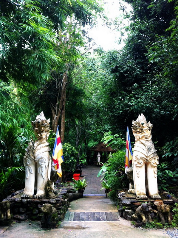 Stone statues of mythical creatures beside a lush green pathway with flags on each side at Pha Lad Temple.