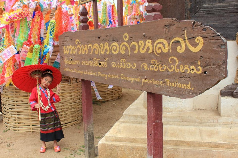 Traditional Thai Dress and Sign at Wat Phan Tao Young girl in traditional Thai attire holding a red umbrella beside a wooden sign at Wat Phan Tao.