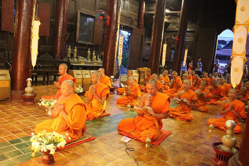 Young monks in orange robes praying inside a wooden temple with books in hand at Wat Phan Tao.