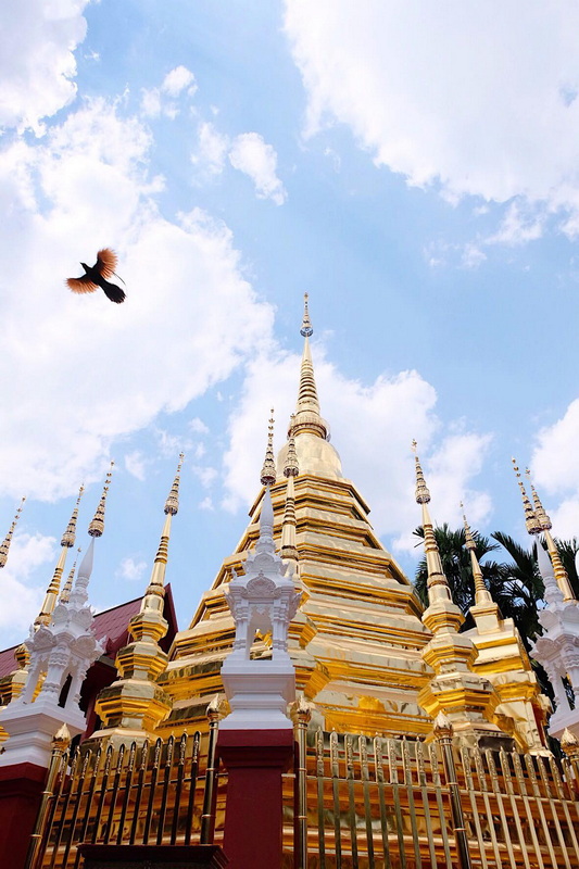 Tall golden and white Buddhist stupa with spires against a blue sky with clouds at Phan Tao Temple.