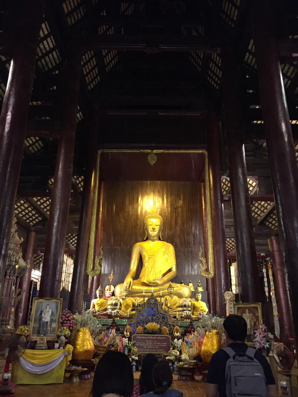 Golden Buddha statue inside a wooden temple with smaller statues and offerings at Wat Phan Tao.