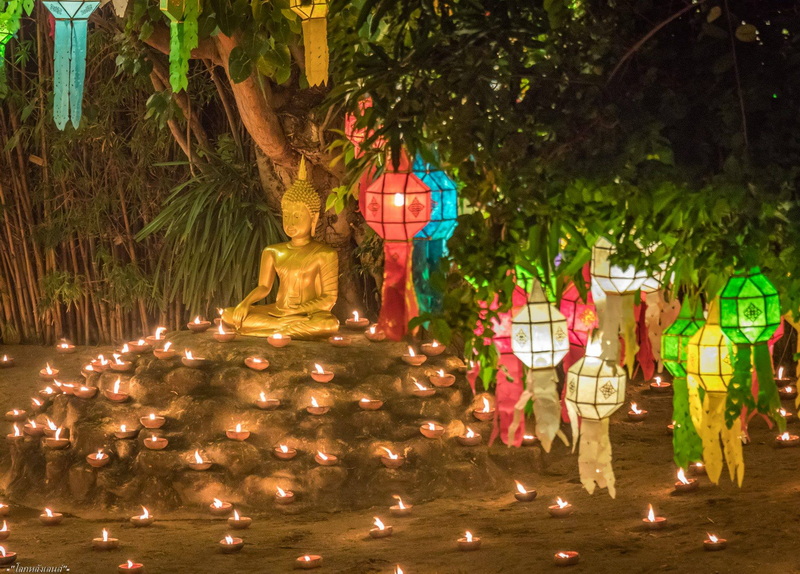 Golden Buddha statue surrounded by candles and hanging lanterns in a lush outdoor setting at Phan Tao Temple.