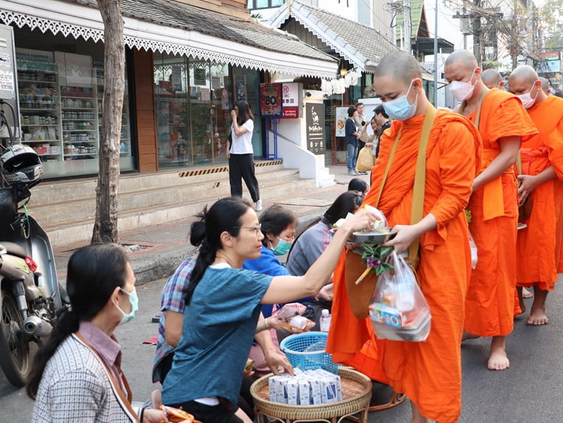 Monks Receiving Alms at Wat Phra Singh Monks receiving alms from people on a street sidewalk with storefronts behind them at Wat Phra Singh.