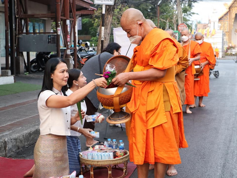 Woman offering a flower to a monk in orange robes during almsgiving on a street at Wat Phra Singh.