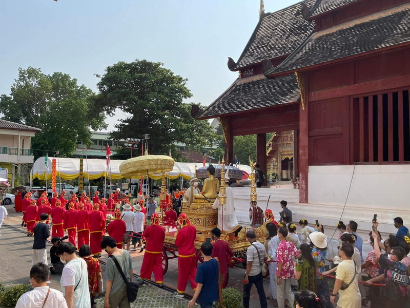 People participating in a cultural procession near a traditional wooden temple with a golden statue on a decorated platform at Wat Phra Singh.