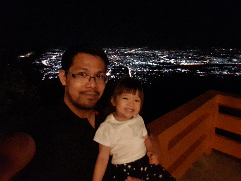 Man and girl smiling on balcony at night with city lights behind them at Doi Suthep temple.