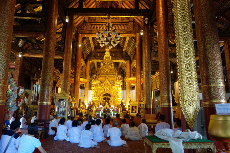People in white sitting inside a gold-adorned temple with wooden pillars and statues at Wat Phra That Sri Chom Thong.