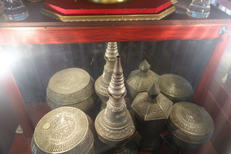 Six metal ritual containers and a tall decorative object inside a glass display case at Wat Phra That Si Chom Thong.