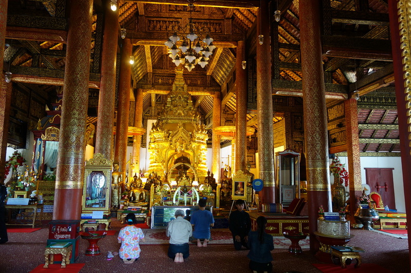 Interior of a richly decorated temple with a golden Buddha statue and praying visitors at Wat Phra That Si Chom Thong.