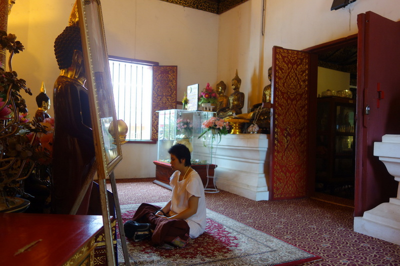 Person sitting on a carpet surrounded by Buddhist statues and decorations inside a temple room at Wat Phra That Sri Chom Thong.