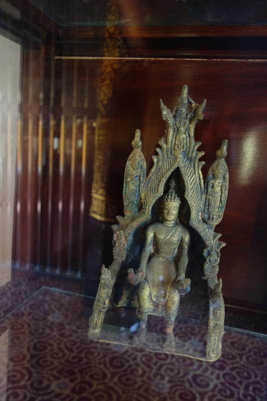 Bronze statue of a standing figure with an ornate arch behind, displayed inside a glass case at Wat Phra That Sri Chom Thong.