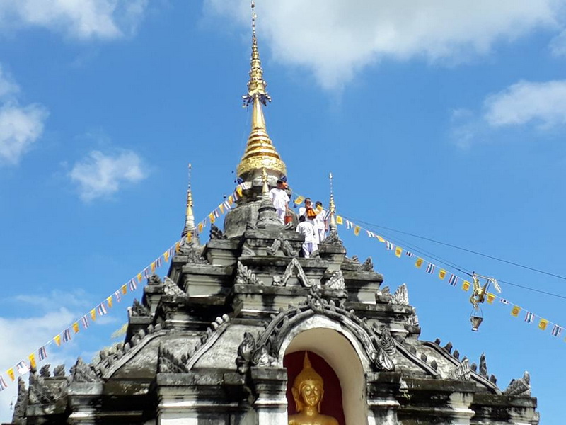 People on a temple roof with golden spire and decorations under blue sky at Wat Phra Yuen.