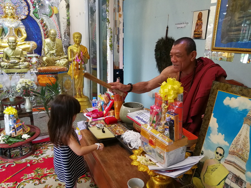 Monk blessing a girl with a wooden stick inside a room with Buddhist statues and photos at Wat Sri Suphan.