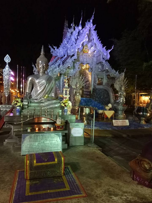 Silver Buddha statue in front of an ornate temple at night with lighting and decorations at Sri Suphan Temple.