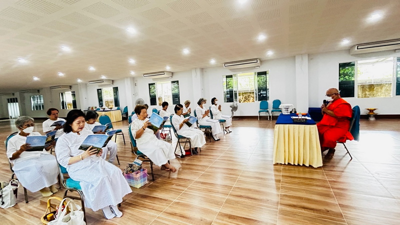 People in white attire listening to a speaker in a bright indoor room at Wat Suan Dok.