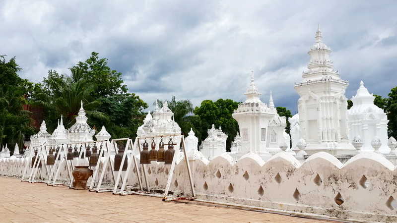 White chedis and bell-shaped structures with trees under cloudy sky at Wat Suan Dok.