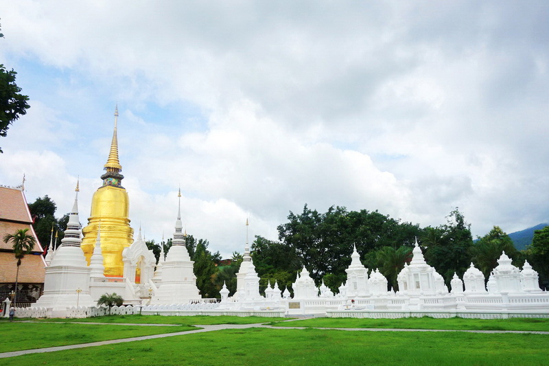 View of a white temple complex with a golden stupa and surrounding smaller stupas, under a cloudy sky at Suan Dok Temple.
