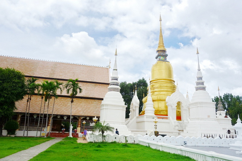 White and gold Buddhist stupa with smaller surrounding stupas under cloudy sky at Suan Dok Temple.
