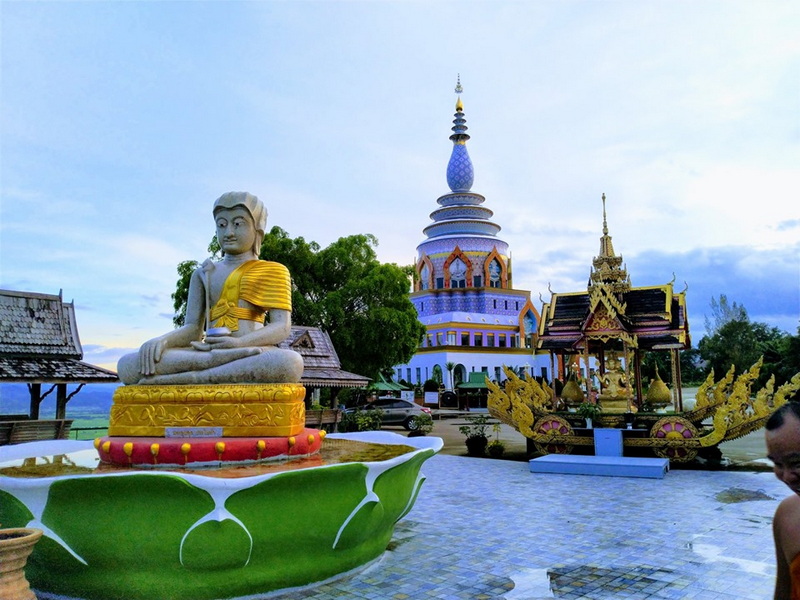 Wat Thaton Temple Scene White Buddha statue with golden base in front of a blue temple spire and ornate structures at Wat Thaton.