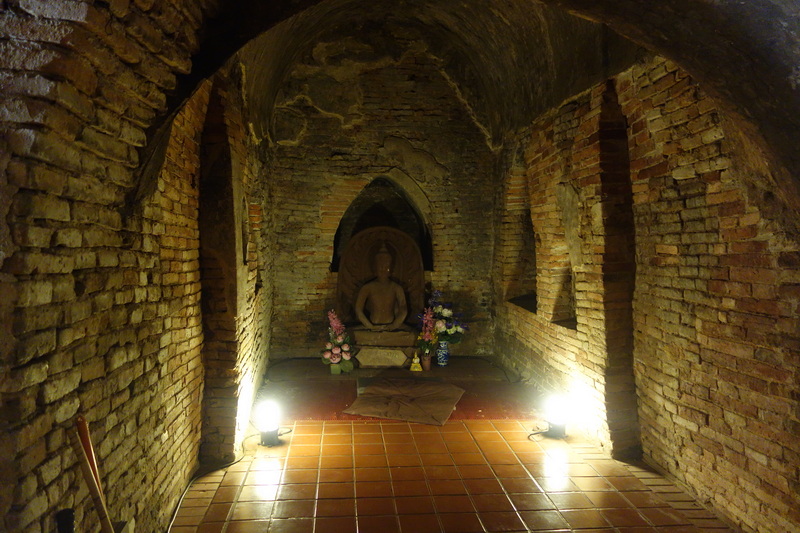 Brick chamber with seated Buddha statue and flower arrangements, illuminated by lights at Wat Umong.