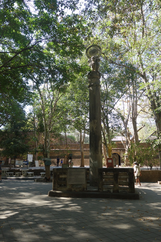 Wooden pole with decorative top in a shaded outdoor space among trees at Wat Umong.
