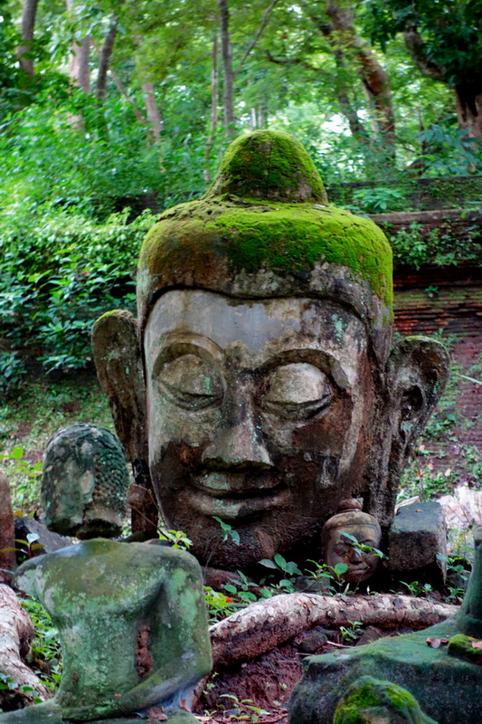 Large moss-covered stone Buddha head with smaller statues in a green forest at Umong Temple.