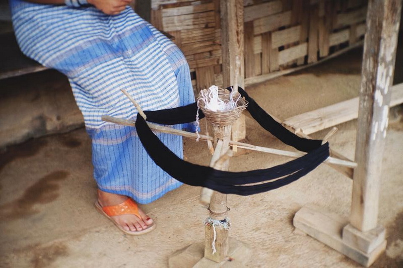 Person sitting on bench with traditional loom and fabric in wooden indoor setting at Mae Hong Son.