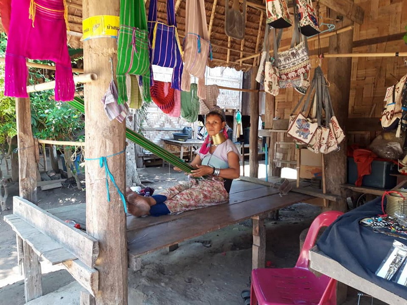 Woman sitting on a wooden platform inside a rustic shop with hanging bags and fabric at Mae Hong Son.