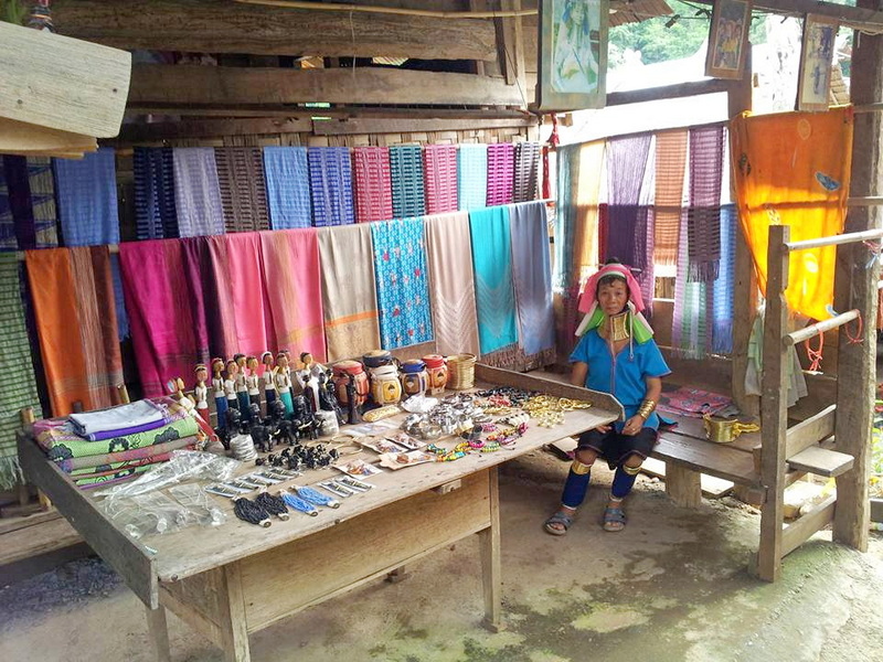 Local Craft Shop Scene at Ban Nam Phiang Din Young girl in traditional attire sitting beside a table of colorful handmade crafts inside a wooden shop at Ban Nam Phiang Din.