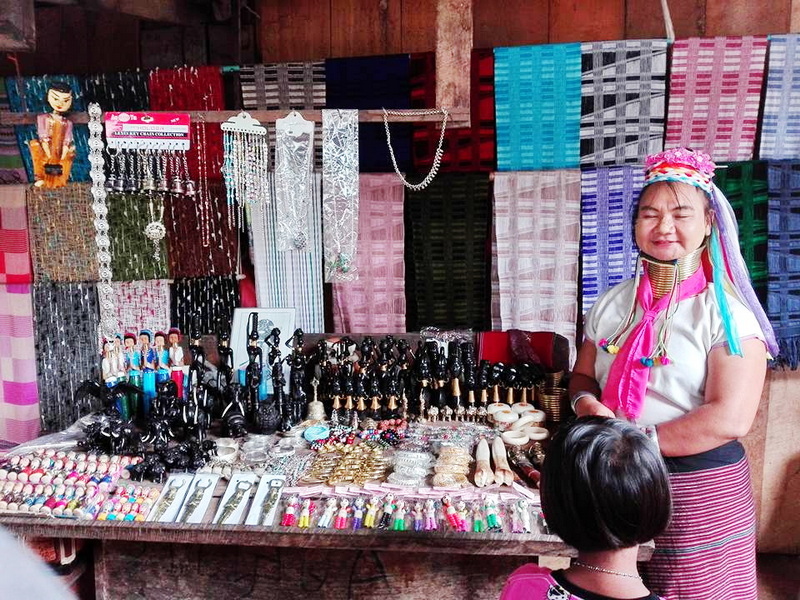 Woman with colorful accessories standing at a jewelry and textile stall displaying various items at Ban Nam Phiang Din.