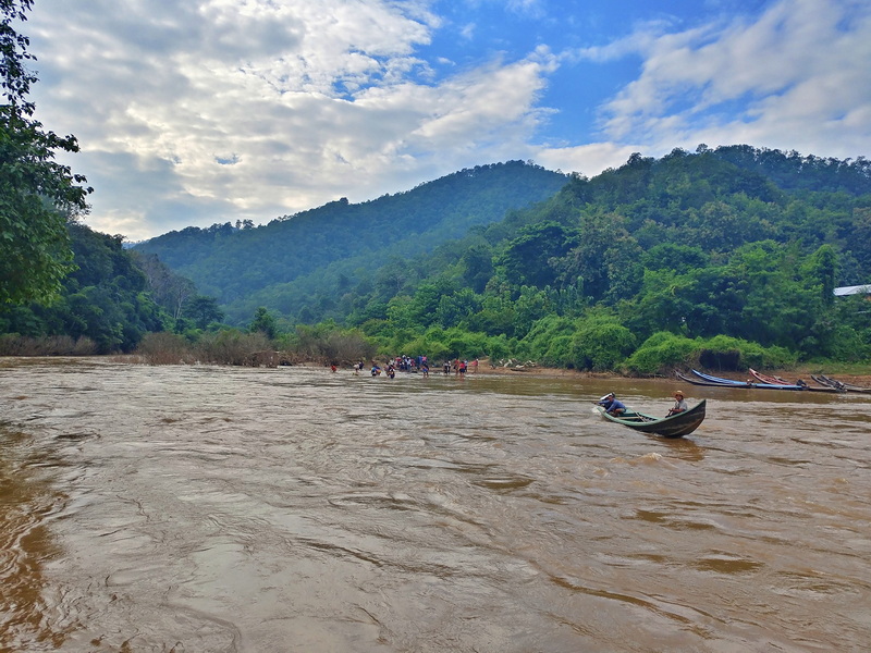 People crossing a river with a boat amidst green forested hills under cloudy sky at Ban Nam Phiang Din.
