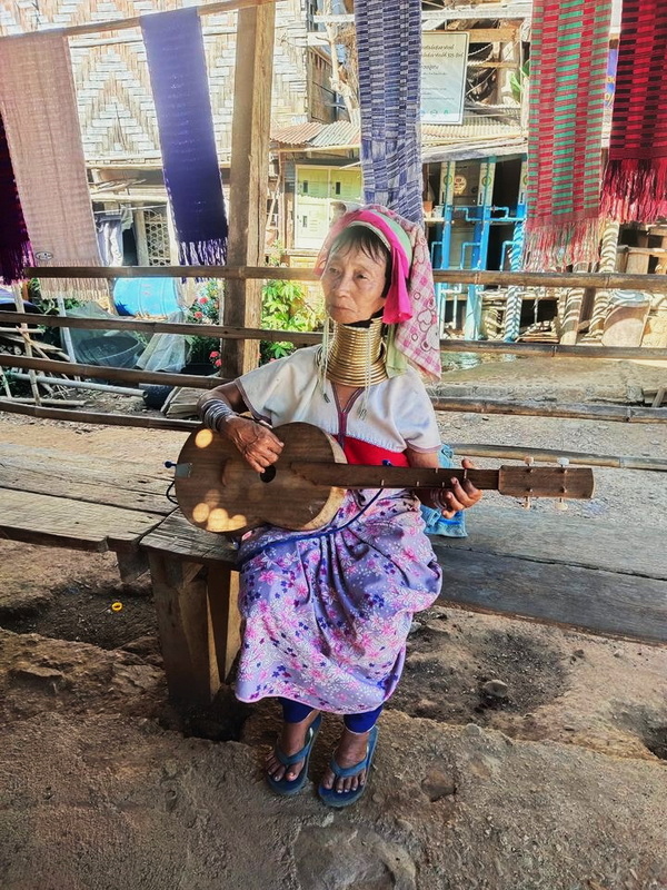 Traditional Musician in Village at Mae Hong Son Elderly woman with brass rings playing guitar in a village with hanging colorful textiles at Mae Hong Son.