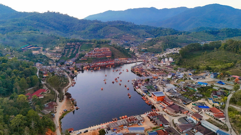 Aerial view of a lake with boats, surrounded by buildings and lush green hills at Ban Rak Thai.