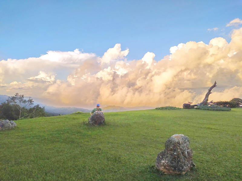 Person sitting on a rock in a green grassy area with clouds and a fallen tree in the background at Huai Nam Dang National Park.