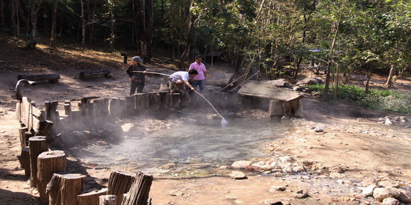 Natural Hot Spring Experience at Huai Nam Dang National Park People soaking in a steaming natural hot spring with wooden logs around and trees nearby at Huai Nam Dang National Park.