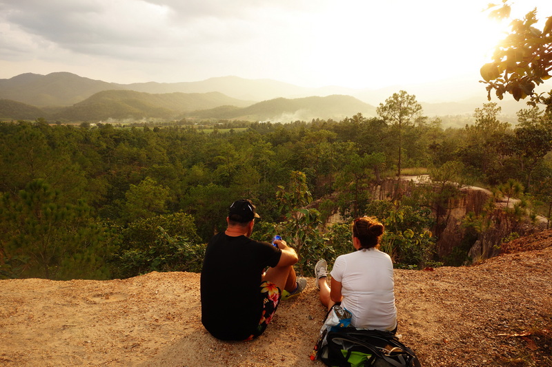 Two people sitting on a cliff edge with a forest and mountains in the background at sunset at Pai Canyon.