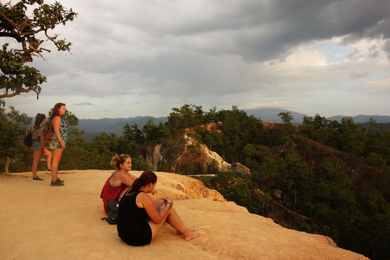 Scenic Hilltop View at Pai Canyon Four people seated and standing on a hilltop overlooking mountains under cloudy sky at Pai Canyon.