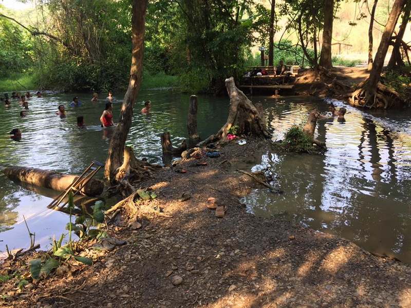 People enjoying a natural hot spring surrounded by lush trees and rocks at Sai Ngam Hot Spring.
