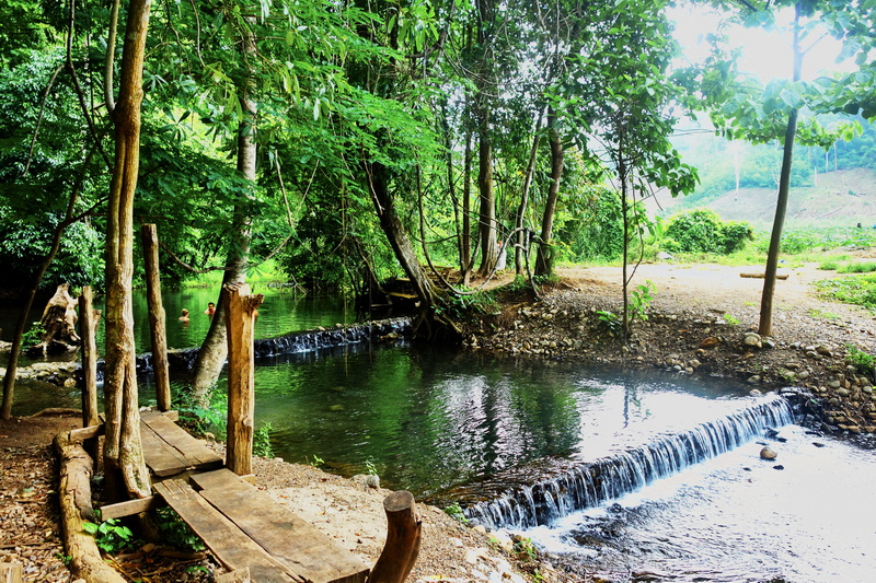 Quiet river with small waterfall surrounded by lush green trees at Sai Ngam Hot Spring.