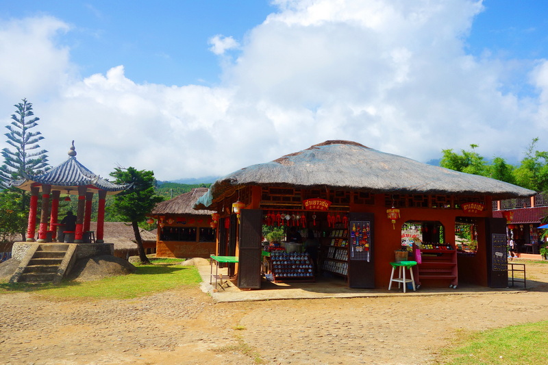 Small souvenir shop with thatched roof and pagoda in the background under a partly cloudy sky at Santichon Village.