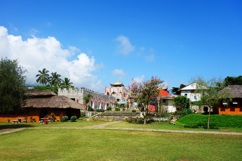 Traditional buildings surrounded by trees and grass under a bright blue sky at Santichon Village.