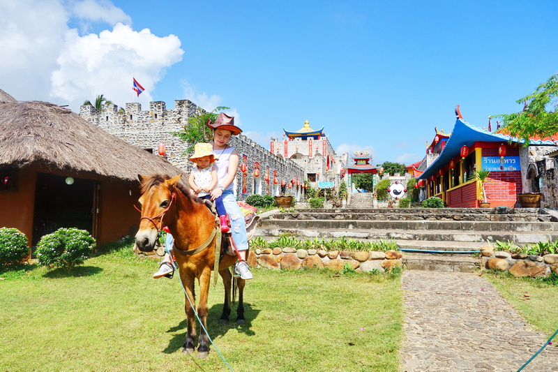 Woman rides a pony with a child in front of colorful structures and castle wall at Santichon Village.